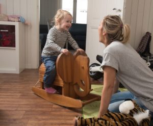 Liten jente på gyngehest smiler mot en voksen som sitter på gulvet