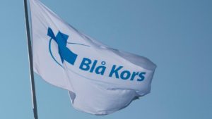 Et hvitt flagg med Blå Kors-logoen mot en blå himmel.