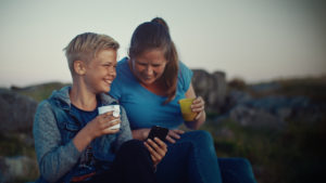 En voksen dame og en ung gutt, mor og sønn, sitter ute i naturen og drikker fra plastkopper og ser på en mobiltelefon.