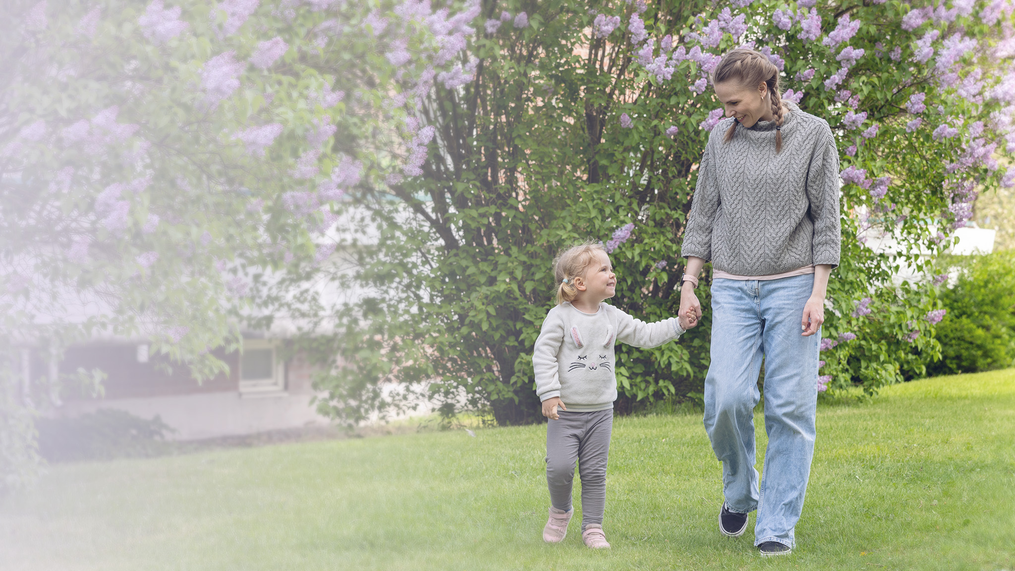 Blå Kors frivillig er tidgiver. Ved å gi av din tid kan du hjelpe andre. En dame leier hender med en liten jente på gressplen med lilla blomster i bakgrunnen.