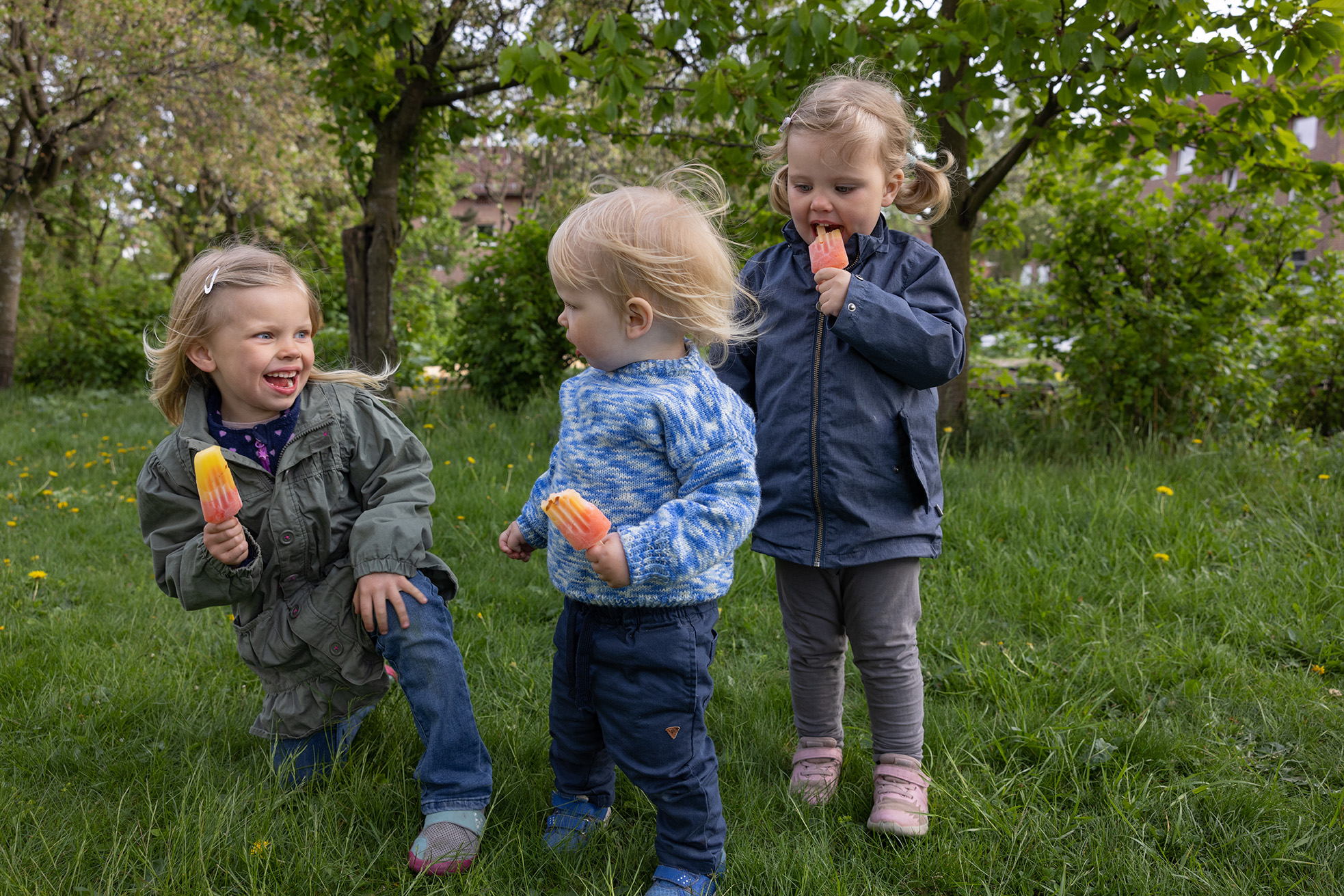 Tre barn som smiler og spiser is ute en fin sommerdag. De er i en grønn park med store trer i bakgrunnen