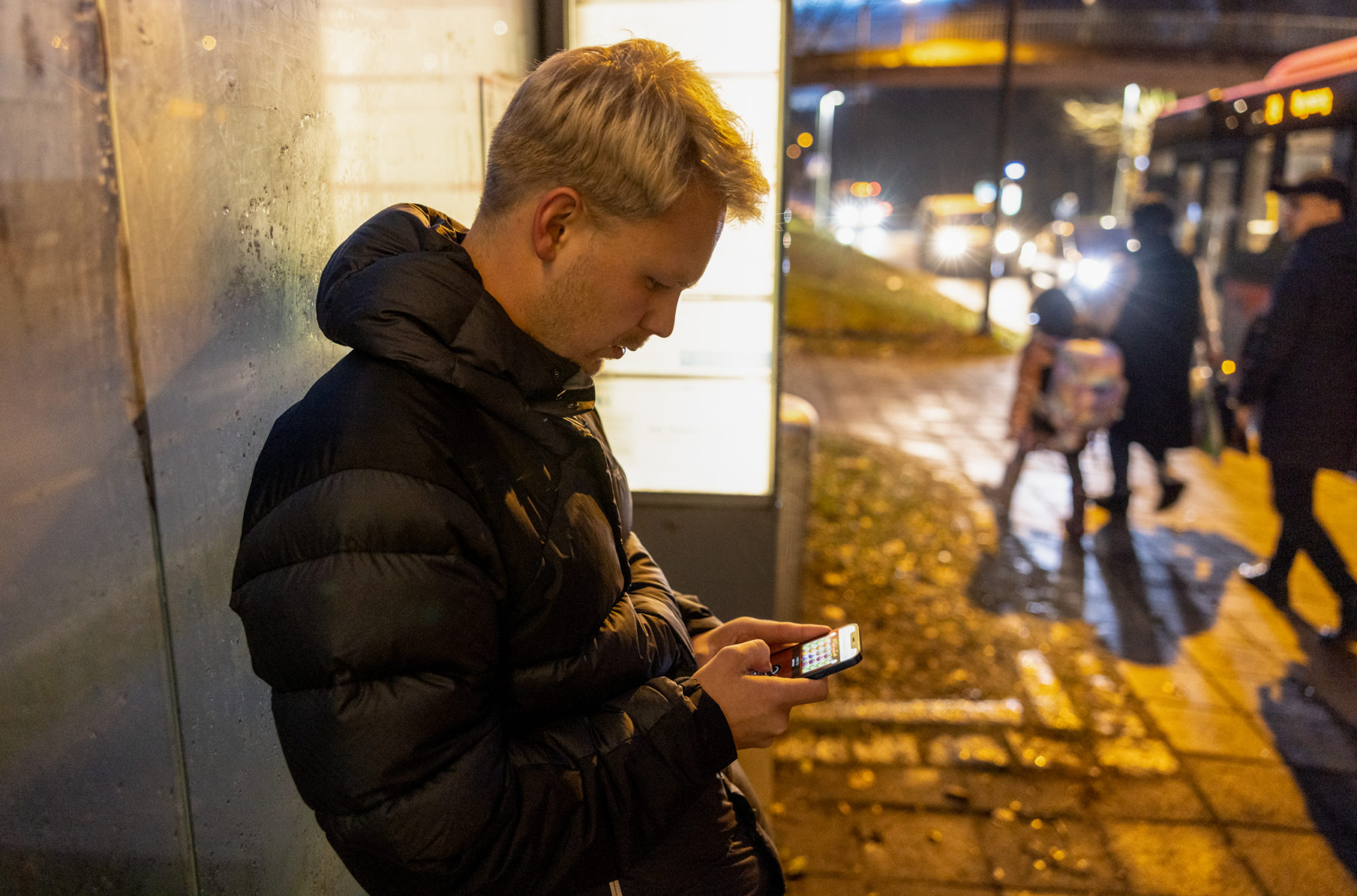En mann spiller konsentrert på mobilen, mens han står og venter på bussholdeplassen.