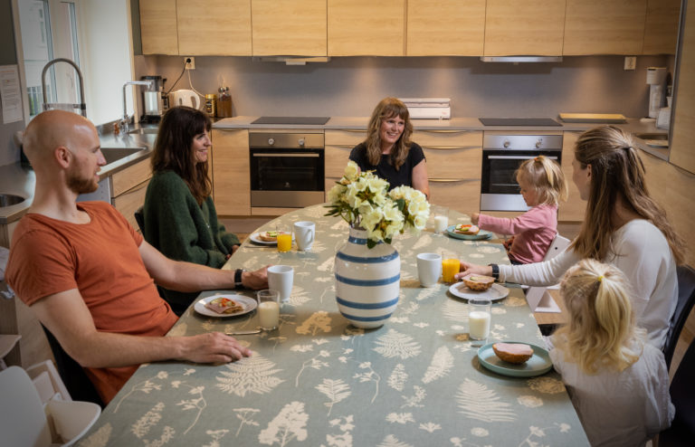 Fire voksne og to barn sitter rundt et spisebord og spiser. Det er en blomsterbukett midt på bordet. Det er hyggelig stemning