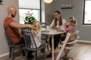 To voksne og to barn sitter rundt et kjøkkenbord på kjøkkenet. Det ene barnet sitter på en barnestol. Det er hyggelig stemning